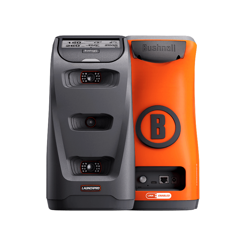 Two golf launch monitors, one black with "LAUNCH PRO," one orange with "Bushnell" and a "B" logo. They stand side-by-side.